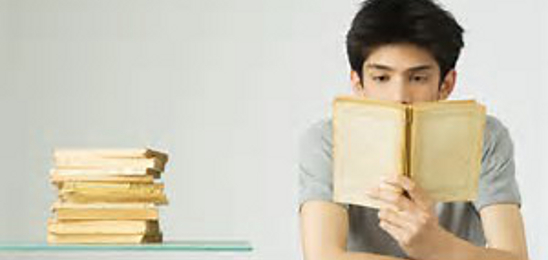 Young man reading a book at a desk. Stack of books on desk.